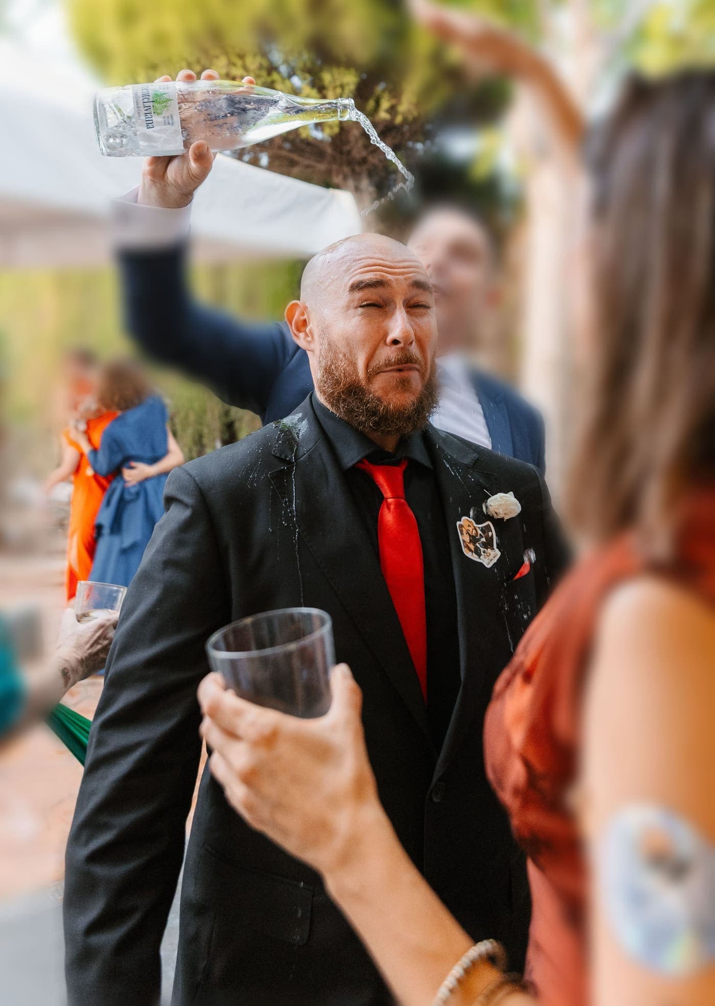 Un novio con traje y corbata roja pone cara de susto mientras un amigo le vacía una botella de agua por encima de la cabeza